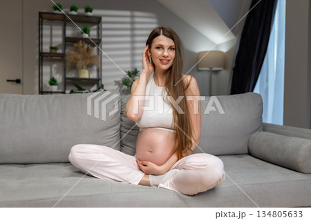 Young expectant mother sits comfortably on a couch, cradling her belly with a gentle smile, anticipating new life Young expectant mother sits comfortably on a couch, cradling her belly with a gentle smile, anticipating new life 134805633