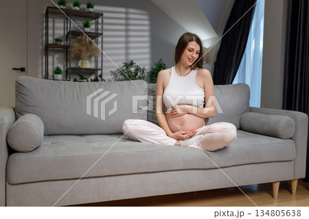 Smiling pregnant woman sits cross-legged on a grey sofa, cradling her belly with her hands, indoors Smiling pregnant woman sits cross-legged on a grey sofa, cradling her belly with her hands, indoors 134805638