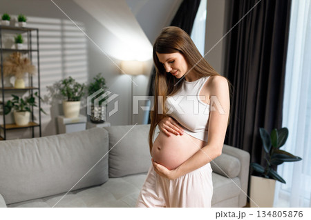 Young woman cradles her pregnant belly with a gentle smile, standing in a softly lit living room with plants and a sofa 134805876
