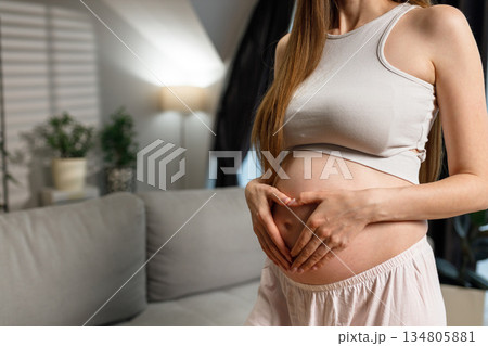 Pregnant woman cradles her belly forming a heart shape with her hands, indoors with soft lighting and home decor elements visible Pregnant woman cradles her belly forming a heart shape with her hands, indoors with soft lighting and home decor elements visible 134805881