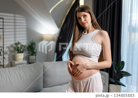Young expectant mother cradles her belly forming a heart shape with her hands, indoors with soft lighting and home decor 134805883