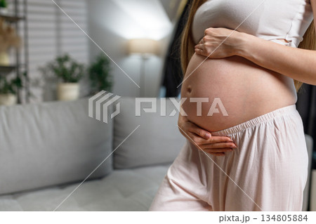 Pregnant woman in light pajamas gently cradles her belly while standing indoors near a sofa and plants Pregnant woman in light pajamas gently cradles her belly while standing indoors near a sofa and plants 134805884