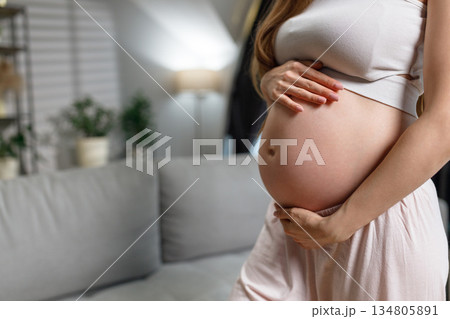 Close-up of pregnant woman's belly with hands cradling it, indoors with soft lighting and blurred background Close-up of pregnant woman's belly with hands cradling it, indoors with soft lighting and blurred background 134805891