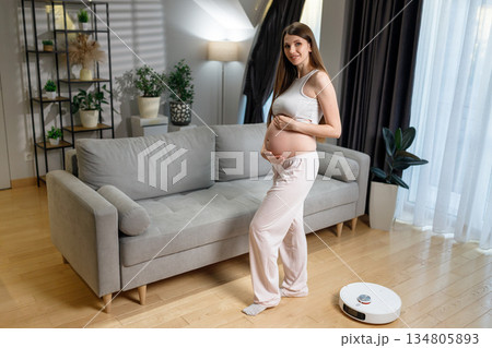 Pregnant woman stands in living room next to sofa with robot vacuum cleaner on wooden floor Pregnant woman stands in living room next to sofa with robot vacuum cleaner on wooden floor 134805893