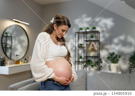 Young woman cradles her pregnant belly with gentle hands, looking down with a serene expression in a cozy living room setting Young woman cradles her pregnant belly with gentle hands, looking down with a serene expression in a cozy living room setting 134805910