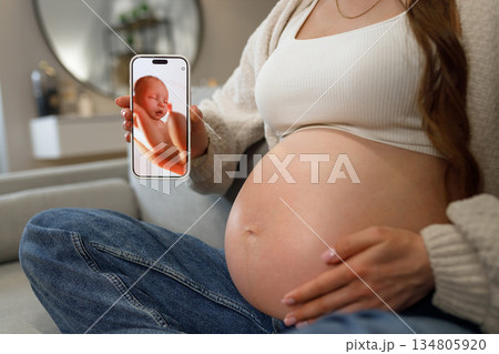 Pregnant woman holding smartphone displaying ultrasound image of her unborn baby while sitting on sofa at home 134805920