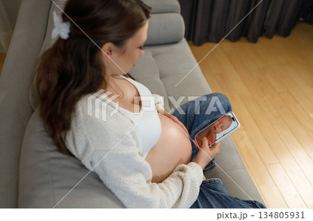 Pregnant woman sits on sofa holding smartphone displaying ultrasound image of her unborn baby, anticipating motherhood Pregnant woman sits on sofa holding smartphone displaying ultrasound image of her unborn baby, anticipating motherhood 134805931