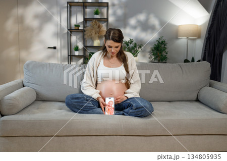 Pregnant woman sits on couch looking at phone displaying ultrasound image, showing anticipation and connection to unborn baby 134805935