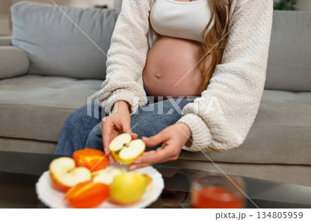 Pregnant woman sitting on sofa holding apple slices and persimmon slices, healthy eating during pregnancy 134805959