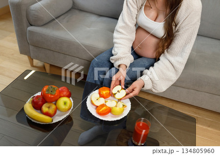 Pregnant woman sits on sofa holding sliced apple and persimmon from plate, healthy snack during pregnancy 134805960
