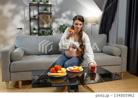 Pregnant woman sits on sofa, eating orange slice and holding glass of juice, with plates of fruit nearby Pregnant woman sits on sofa, eating orange slice and holding glass of juice, with plates of fruit nearby 134805962
