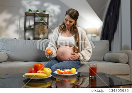 Expecting mother sits on couch holding persimmon slice, healthy food on table, indoor setting, warm lighting 134805986