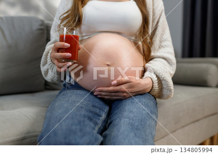 Third trimester pregnant woman sits on sofa holding glass of tomato juice, cradling her belly with her hands 134805994