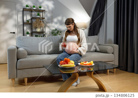 Pregnant woman sits on sofa holding glass of juice and touching belly, fruit on coffee table Pregnant woman sits on sofa holding glass of juice and touching belly, fruit on coffee table 134806125