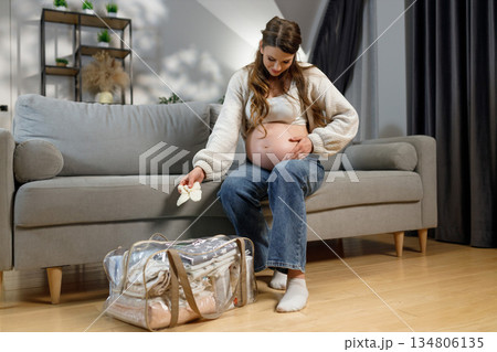 Expecting mother sits on sofa, holding baby clothes, preparing hospital bag for childbirth, feeling anticipation and readiness 134806135