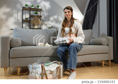 Expecting mother sits on sofa preparing baby items for hospital bag, smiling gently at camera 134806143
