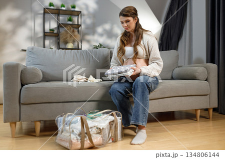 Expecting mother sits on sofa folding baby garments preparing hospital bag for childbirth Expecting mother sits on sofa folding baby garments preparing hospital bag for childbirth 134806144