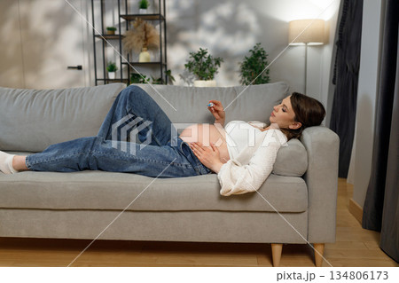 Pregnant woman resting on a sofa, holding a small object in her hand, with her belly exposed and hands resting on it Pregnant woman resting on a sofa, holding a small object in her hand, with her belly exposed and hands resting on it 134806173