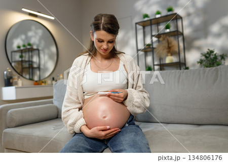 Expecting mother sits on sofa holding thermometer near her belly, checking temperature during pregnancy Expecting mother sits on sofa holding thermometer near her belly, checking temperature during pregnancy 134806176