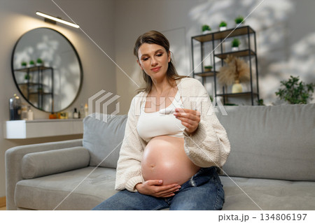 Pregnant woman with large belly sits on sofa holding pregnancy test kit, looking at it with anticipation and joy Pregnant woman with large belly sits on sofa holding pregnancy test kit, looking at it with anticipation and joy 134806197