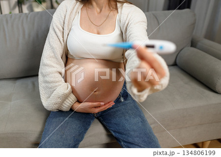 Pregnant woman sitting on sofa holding thermometer, checking temperature during pregnancy Pregnant woman sitting on sofa holding thermometer, checking temperature during pregnancy 134806199