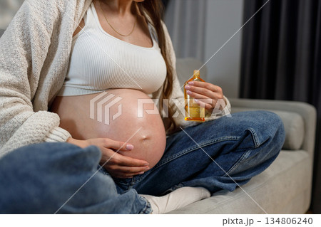 Pregnant woman holding bottle of oil for belly care while sitting on couch, expecting mother nurturing skin Pregnant woman holding bottle of oil for belly care while sitting on couch, expecting mother nurturing skin 134806240
