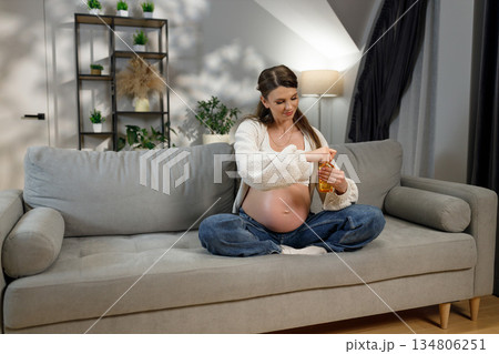 Pregnant woman sitting cross-legged on sofa applying body oil to her belly for stretch mark prevention 134806251