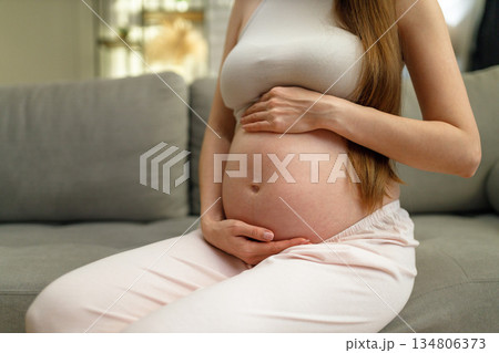 Pregnant woman sits on sofa, cradling her belly with both hands, showing anticipation and maternal love 134806373