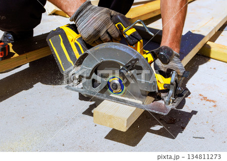 Worker cuts wooden beams board using circular saw while standing on concrete surface. Worker cuts wooden beams board using circular saw while standing on concrete surface. 134811273