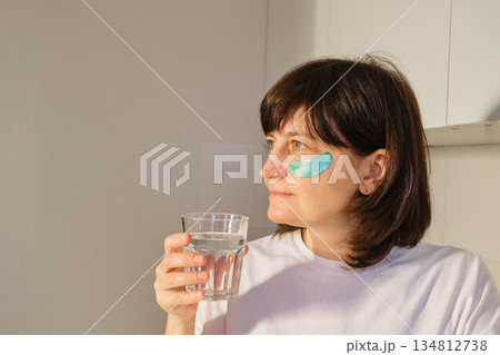 Mature Woman applying under eye patches and drinking water for morning self-care and wellness 134812738