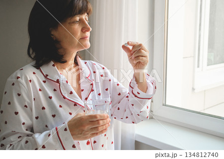 Mature Woman in pajamas holding pill and glass of water for daily treatment Mature Woman in pajamas holding pill and glass of water for daily treatment 134812740