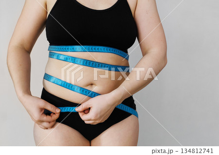 Woman standing against plain background measuring her waist with blue tape measure 134812741