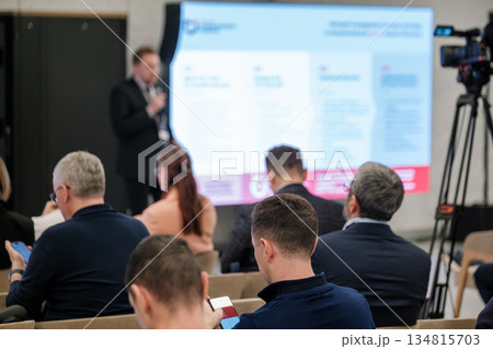 Business conference in progress with speaker presenting to seated audience and camera crew 134815703
