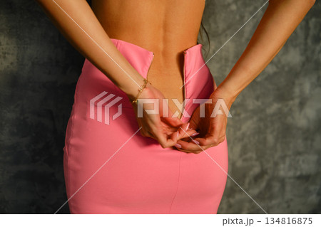 Woman adjusts zipper on pink skirt against grey wall in indoor setting during daytime 134816875