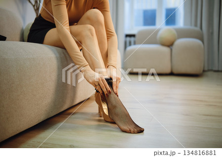 Unrecognizable woman sitting on couch putting on black fishnet stockings in modern room during daytime Unrecognizable woman sitting on couch putting on black fishnet stockings in modern room during daytime 134816881