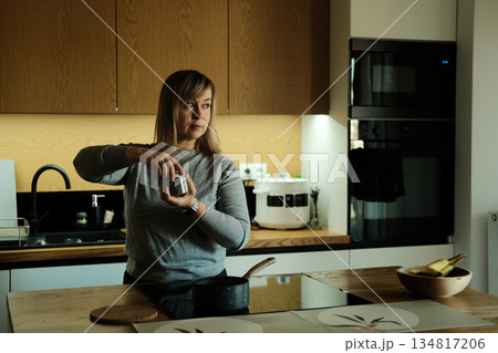 Woman stands in modern kitchen holding jar of herbs while looking away from camera. Natural flavor enhancers, plant-based nutrition, holistic cooking, wellness kitchen. 134817206