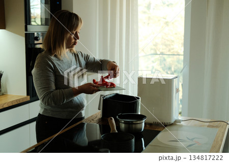 Woman adds sliced bell pepper to air fryer in kitchen. Nutritious food, simple recipes, culinary wellness, healthy meal prep. Woman adds sliced bell pepper to air fryer in kitchen. Nutritious food, simple recipes, culinary wellness, healthy meal prep. 134817222