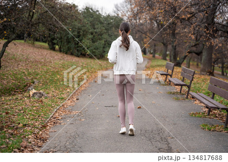 Woman jogging in autumn park wearing white hoodie. Running along path in peaceful city park, showcasing active lifestyle and self care outdoors. 134817688