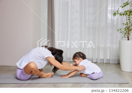 Mother helping young daughter stretching on a yoga mat during home workout 134822206