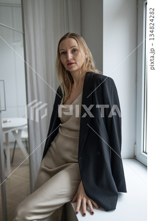 Elegant woman in beige dress and black blazer sitting by window in minimalist interior Elegant woman in beige dress and black blazer sitting by window in minimalist interior 134824282
