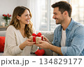 young couple at home celebrating Valentine's Day, smiling naturally, exchanging a small wrapped gift with a red ribbon 134829177