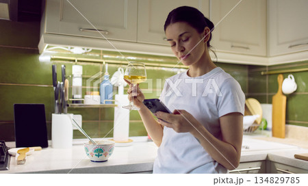 Woman holding glass of wine and checking smartphone in cozy home kitchen. 134829785