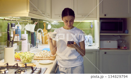 Woman checking smartphone while holding cup in cozy home kitchen. 134829794