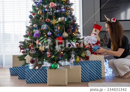 Young woman placing wrapped gifts under decorated Christmas tree with colorful ornaments. Young woman placing wrapped gifts under decorated Christmas tree with colorful ornaments. 134831342