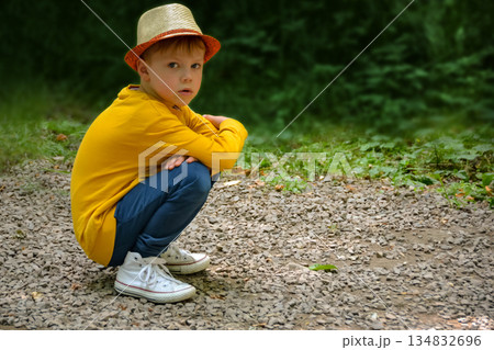 A boy wearing a yellow shirt and a hat is kneeling on a gravel road. High quality photo 134832696