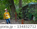 A toddler in a cap and yellow shirt wanders through the forest, surrounded by trees, grass, and terrestrial plants 134832712