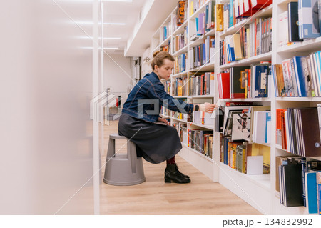 woman is sitting at shelves in library, student is choosing book. library is filled with books. 134832992