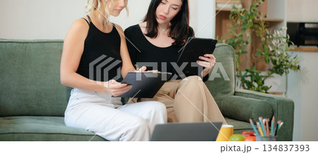 Two women collaborating on a project at home, brainstorming with a laptop and notebook in a cozy setting. 134837393