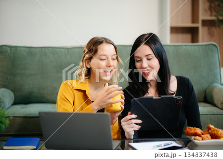 Two women collaborating on a project at home, brainstorming with a laptop and notebook in a cozy setting. Two women collaborating on a project at home, brainstorming with a laptop and notebook in a cozy setting. 134837438