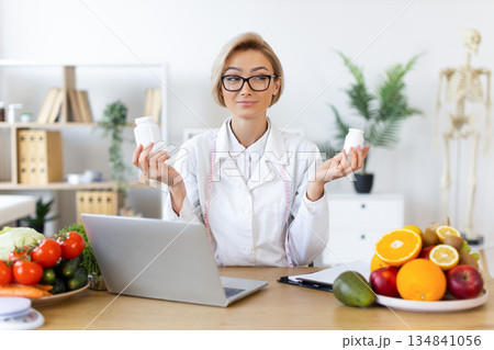 A dietitian holds up two supplement bottles, surrounded by fruits, vegetables, and a laptop, suggesting a healthy lifestyle. A dietitian holds up two supplement bottles, surrounded by fruits, vegetables, and a laptop, suggesting a healthy lifestyle. 134841056